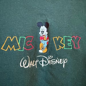 May include: Dark green sweatshirt featuring an embroidered Mickey Mouse design. The word "MICKEY" is embroidered in colorful letters above the character, with "Walt Disney" embroidered below in white.
