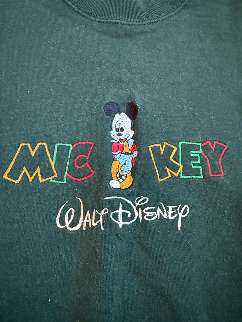 May include: Dark green sweatshirt featuring an embroidered Mickey Mouse design. The word "MICKEY" is embroidered in colorful letters above the character, with "Walt Disney" embroidered below in white.