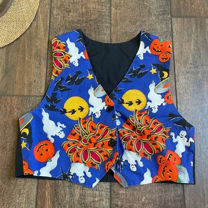 May include: A Halloween-themed vest with a blue background featuring pumpkins, ghosts, bats, and moons. The vest has a black lining and is buttoned up. The vest is a festive garment.