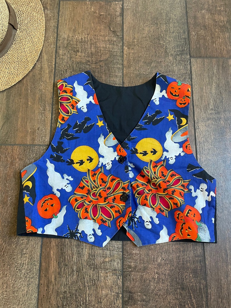 May include: A Halloween-themed vest with a blue background featuring pumpkins, ghosts, bats, and moons. The vest has a black lining and is buttoned up. The vest is a festive garment.