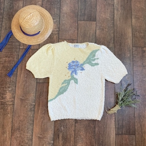 Op de afbeelding: Een crèmekleurige gebreide trui met een bloemenpatroon in tinten blauw, groen en geel. De trui heeft korte mouwen en een ronde hals. Het label "KOET" is zichtbaar op de trui.