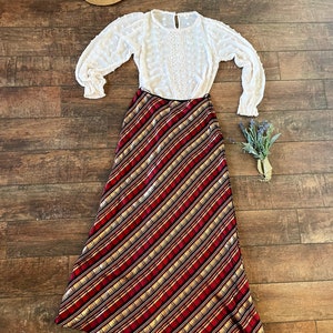 May include: An ensemble featuring a white blouse with lace detailing and long sleeves, paired with a long, diagonally striped skirt in red, gold, and black. A straw hat and a small bouquet of lavender complete the outfit.