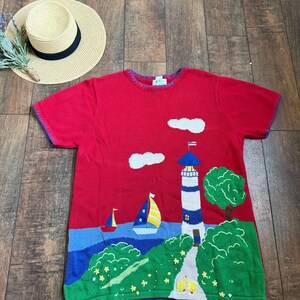 Może przedstawiać: Czerwony sweter z krótkim rękawem z motywem marynistycznym. Sweter przedstawia latarnię morską, żaglówki i zielony krajobraz na tle niebieskiego morza i czerwonego nieba. W tle widoczny jest słomkowy kapelusz z czarną opaską.