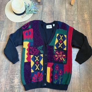 May include: A vintage, button-down cardigan sweater with a patchwork design. The sweater features a V-neck and long sleeves. The color palette includes black, red, yellow, green, and purple. A straw hat and lavender sprig are also in the image.