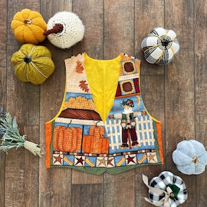 Puede incluir: Un chaleco amarillo con un diseño de patchwork que presenta un espantapájaros, calabazas y estrellas. El chaleco está hecho de tela y tiene una textura acolchada.