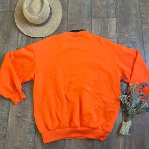 May include: Orange sweatshirt with a black collar and long sleeves. The sweatshirt is laying flat on a wooden surface.