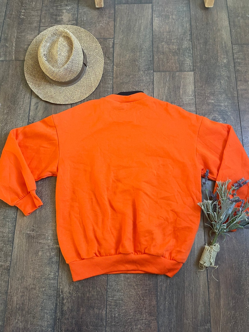 May include: Orange sweatshirt with a black collar and long sleeves. The sweatshirt is laying flat on a wooden surface.