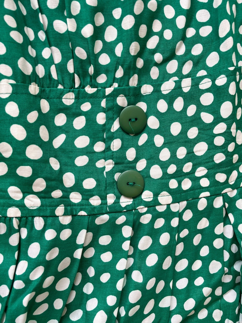 May include: Green fabric with white polka dots and two green buttons. The fabric is pleated and has a seam running horizontally across the middle.