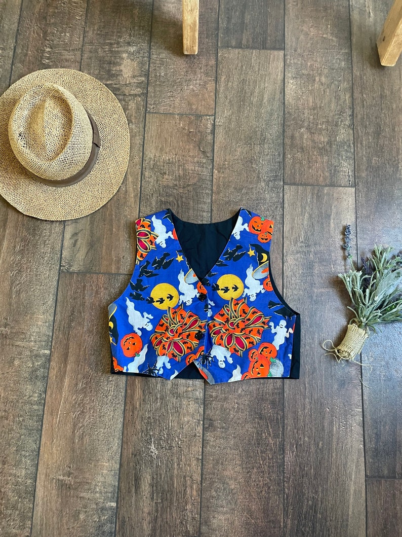 May include: A blue Halloween-themed vest with orange pumpkins, white ghosts, and black bats. The vest has a black lining and is buttoned. A straw hat and dried flowers are also in the image.