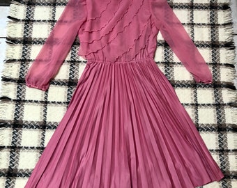 May include: A dusty rose-colored dress with long, sheer sleeves and a pleated skirt. The bodice features layered ruffles across the chest. The dress has a keyhole opening at the back of the neck. A vintage-style garment.
