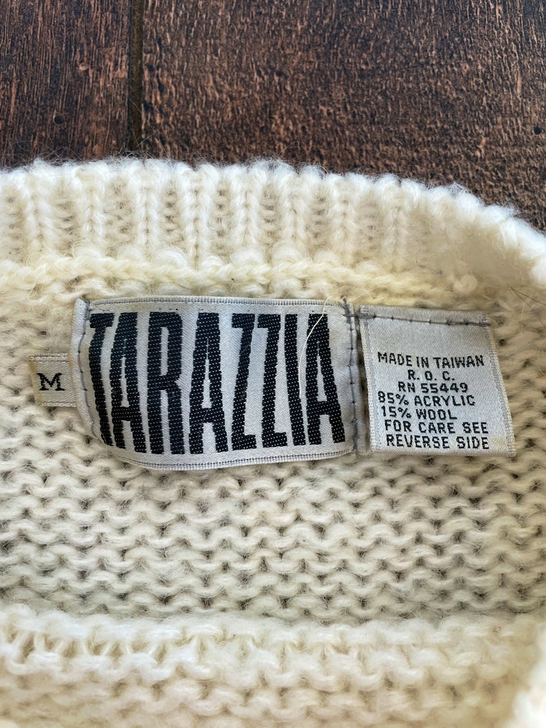 May include: White knitted sweater with a white tag that says "KARAZZIA" and "MADE IN TAIWAN R.O.C. RN 55449 85% ACRYLIC 15% WOOL FOR CARE SEE REVERSE SIDE".