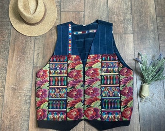 May include: A black waistcoat with a colourful embroidered pattern on the front. The waistcoat has a blue and white striped fabric on the top and a black fabric on the bottom. The waistcoat has a button closure.