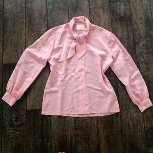 Könnte beinhalten: Eine rosa Bluse mit langen Ärmeln, einem Schleifenkragen und Knöpfen. Die Bluse hat eine lockere Passform und ist aus einem leichten Stoff gefertigt.