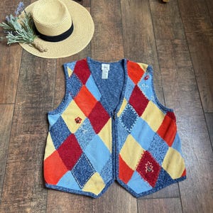 Puede incluir: Un chaleco colorido estilo patchwork con cierre de cremallera. El chaleco presenta un patrón de diamantes en rojo, naranja, amarillo y azul. También se ve un sombrero de paja con una banda negra y una ramita de lavanda.