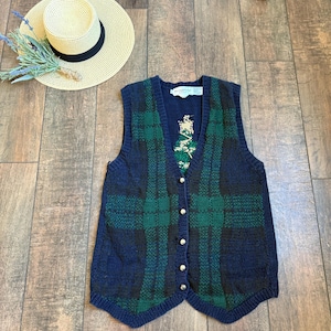 Könnte beinhalten: Eine marineblaue und grüne karierte Strickweste mit silbernen Knöpfen. Ein Strohhut mit schwarzem Band und Lavendelzweigen liegt neben der Weste. Die Weste hat ein kleines gesticktes Design auf der Vorderseite.