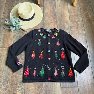 Könnte beinhalten: Schwarze Weihnachtsstrickjacke mit bunten, gestickten Weihnachtsbäumen und Geschenkbox-Akzenten. Die langärmlige Strickjacke hat einen Knopfverschluss. Ein Strohhut und ein Lavendelzweig sind ebenfalls im Bild zu sehen.