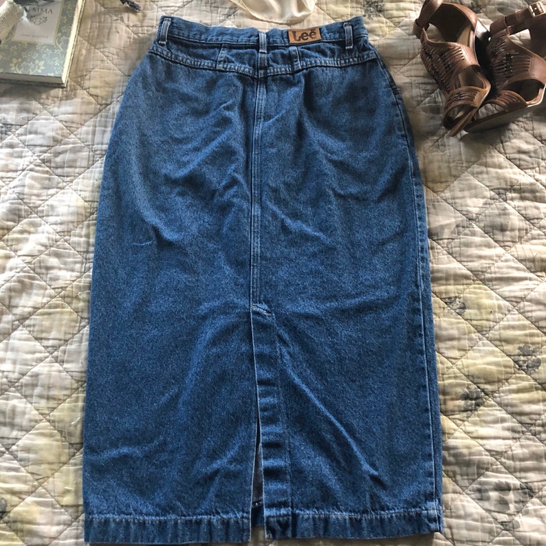 May include: A blue denim skirt with a front slit and a button closure. The skirt has a brown leather label with the word "Lee" on it.