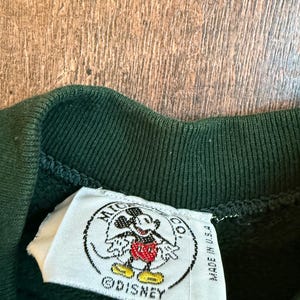 May include: Close-up of a dark green sweatshirt collar with a white tag featuring a Mickey Mouse graphic. The tag reads "MICKEY CO." and "MADE IN U.S.A." with the Disney logo.