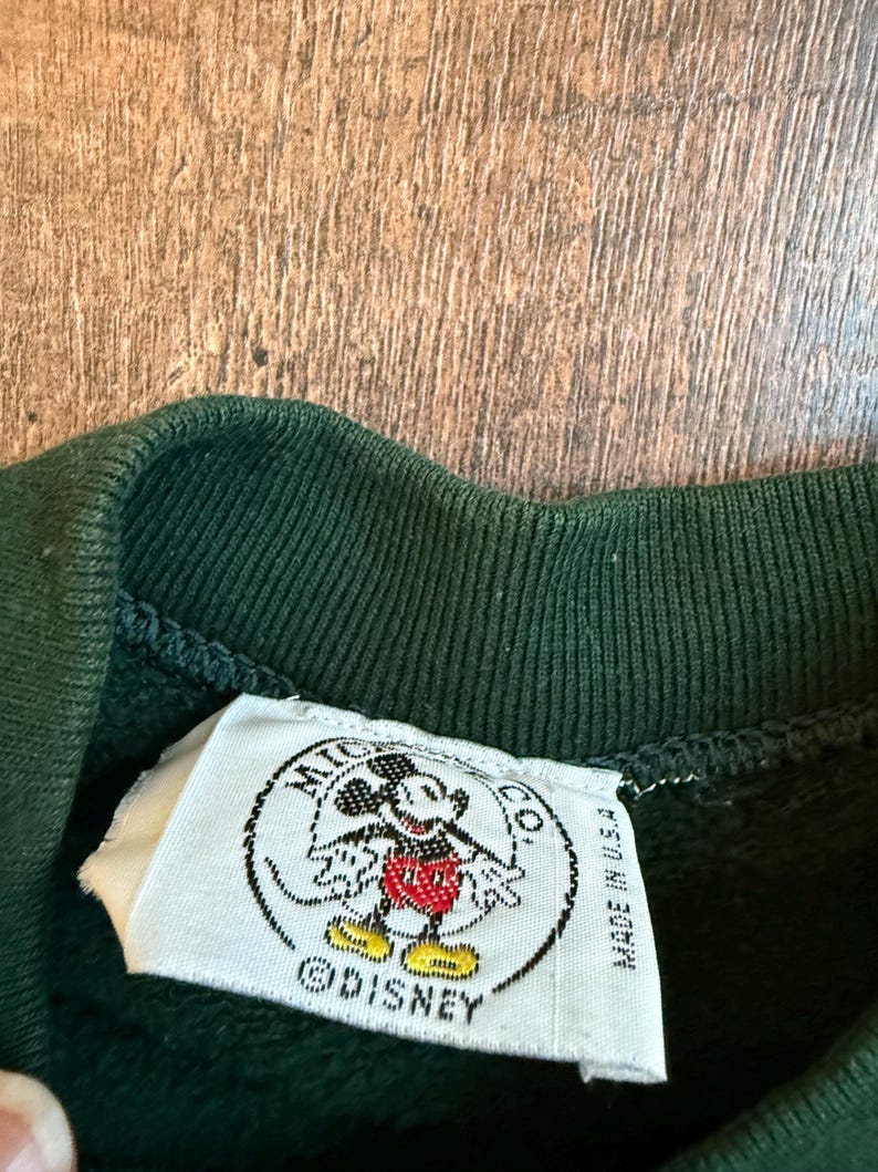 May include: Close-up of a dark green sweatshirt collar with a white tag featuring a Mickey Mouse graphic. The tag reads "MICKEY CO." and "MADE IN U.S.A." with the Disney logo.