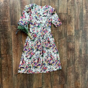 May include: A white floral midi dress with a lace neckline and short sleeves. The dress has a gathered skirt and a fitted bodice. The dress is photographed on a wooden background.