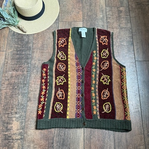 Op de afbeelding: Een veelkleurige gebreide vest met een V-hals en knoopsluiting. Het vest toont bladmotieven en streepjespatronen in herfsttinten van groen, bruin, rood en geel. Een strohoed met een zwarte band is zichtbaar op de achtergrond.