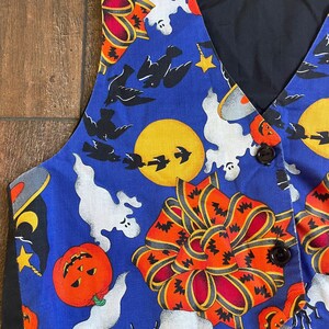 May include: A blue Halloween-themed vest with a black back and trim. The vest features a repeating pattern of orange pumpkins, white ghosts, black bats, and large orange bows. Two brown buttons secure the front.
