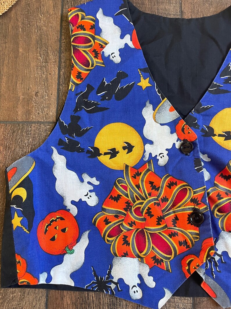 May include: A blue Halloween-themed vest with a black back and trim. The vest features a repeating pattern of orange pumpkins, white ghosts, black bats, and large orange bows. Two brown buttons secure the front.
