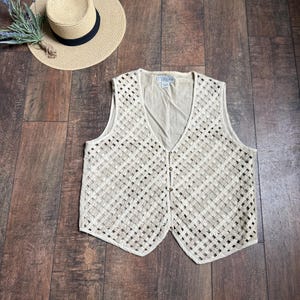 May include: A beige woven vest with a V-neck and button closure. The vest has a lattice design and is paired with a straw hat with a black band. A sprig of lavender is placed near the hat.