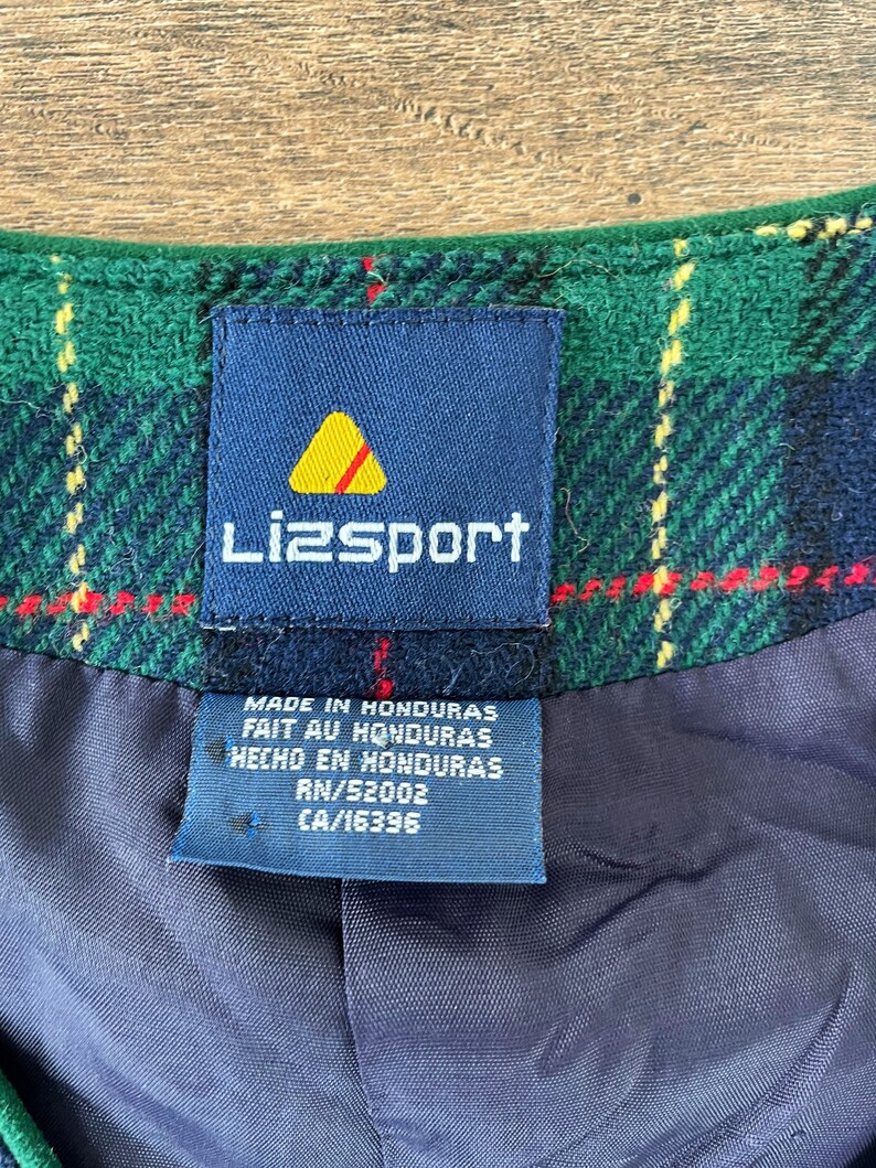 May include: A blue and white Lizsport label with a yellow triangle logo. The label is attached to a green and black plaid fabric. The label reads "Made in Honduras, Fait au Honduras, Hecho en Honduras, RN/52002, CA/16396".