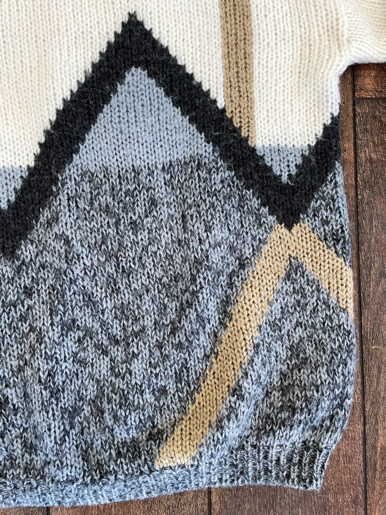 May include: A knitted sweater with a geometric pattern in shades of white, gray, black, and beige.