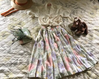 May include: A white blouse with a floral print skirt, a straw hat, and brown heeled sandals. The outfit is laid out on a white quilted bedspread.
