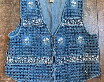 May include: A blue denim vest with a woven design. The vest features floral embroidery and white lace trim. It has a button-up front and a V-neck. The vest is a vintage style.
