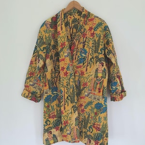 May include: A long-sleeved yellow jacket featuring a vibrant, multi-colored botanical and figurative print. The garment has a wide collar, an open front, and a quilted texture throughout, hanging from a wooden hanger against a plain wall.