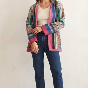 Peut inclure: Une veste à manches longues ouverte sur le devant avec un motif patchwork multicolore composé de rayures et de formes géométriques dans des tons rose, bleu, vert et marron, portée sur un débardeur crème, un jean bleu foncé et des bottines marron.