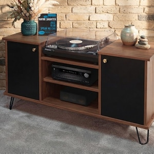 May include: A walnut-toned media console with two black cabinet doors, gold-toned knobs, and black hairpin legs. The top surface holds a record player with a clear cover, a Wayne Shorter vinyl record, a teal vase, and a beige ceramic vase.