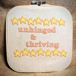 May include: A square embroidery hoop containing a piece of light beige fabric with cross-stitched text that reads unhinged & thriving in a peach color. The text is surrounded by two rows of seven yellow stars, one row above and one row below.