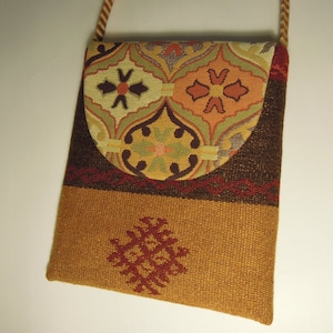 Puede incluir: Un pequeño bolso bandolera marrón y dorado con cierre de solapa. La solapa tiene un tejido estampado de colores con diseños geométricos. El bolso tiene una correa marrón.