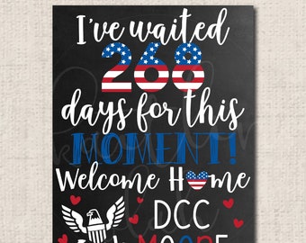 May include: Black chalkboard sign with the text "I've waited 268 days for this MOMENT! Welcome home DCC MOORE" in white. The numbers 268 are decorated with red, white, and blue American flag designs. There are small red hearts and a white eagle with red, white, and blue wings and a blue anchor.