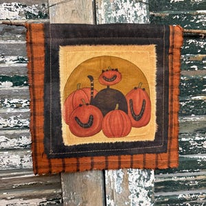 May include: A square fabric wall hanging featuring a central illustration of a black cat with a striped tail sitting among four orange pumpkins with stitched smiles. The piece has layered borders in dark brown and orange plaid fabric with frayed edges.