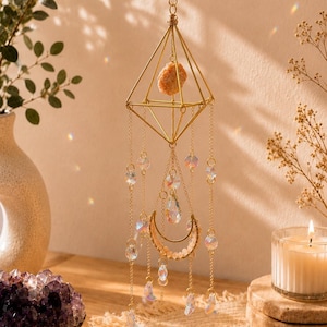 May include: A gold-toned geometric hanging ornament featuring a central diamond-shaped wire frame with a raw peach-colored stone inside. The piece includes multiple dangling gold chains adorned with clear iridescent crystal beads and a crescent moon charm.