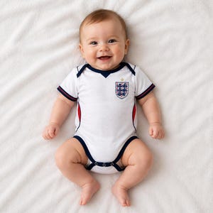 Könnte beinhalten: Ein weißer Baby-Body mit kurzen Ärmeln, marineblauen und roten Abschlüssen, einem marineblauen V-Ausschnitt und einem Wappen auf der Brust, das drei Löwen mit einem kleinen goldenen Stern darüber zeigt. Das Kleidungsstück hat marineblaue Paspeln.