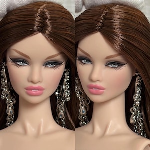 May include: A fashion doll with long, straight brown hair, light skin, and painted facial features including winged eyeliner and pink lips. The doll wears long, dangling silver-toned earrings featuring multiple chain links and small blue accents.