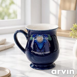 Puede incluir: Una taza de cerámica azul marino profundo con acabado brillante, que presenta un motivo floral estilizado central en tonos azules, verdes y dorados. La taza tiene un cuerpo redondeado, un borde acampanado, un asa resistente y una base.