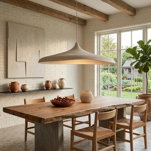 May include: A large, wide, beige pendant light fitting with a smooth, curved shape hangs above a rectangular wooden dining table with a live edge. The table is supported by two grey concrete pedestals and surrounded by four wooden chairs.