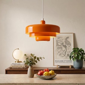 May include: A bright orange pendant light with a three-tiered, layered circular shade design hangs from a white cord. The fixture features a smooth, glossy finish and emits a warm glow from the open bottom, illuminating the space below.