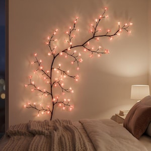 Op de afbeelding: Een decoratieve wandlamp in de vorm van een tak met donkerbruine stelen, versierd met talloze kleine, oplichtende roze LED-bloemen. De tak strekt zich uit over de muur boven een bed en zorgt voor een zachte, sfeervolle verlichting.