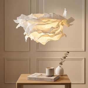 May include: A white pendant light fixture with a layered, ruffled, paper-like shade that emits a warm glow. The shade features irregular, organic folds and curves, suspended from a thin cord, creating a soft, diffused lighting effect.