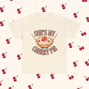 Puede incluir: Una camiseta de manga corta color crema con un gráfico central de una tarta de cerezas con nata montada y una cereza encima. El texto "SHE'S MY CHERRY PIE" está impreso en letras verdes y rojas sobre y debajo de la ilustración de la tarta.