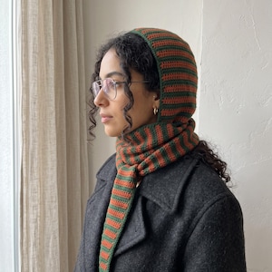 May include: A crochet hood and scarf combination featuring alternating horizontal stripes in dark green and burnt orange. The headpiece covers the top of the head and extends into a long, pointed scarf that drapes down the front of the chest.