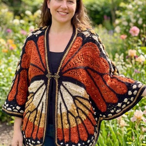 Può includere: Un cardigan all'uncinetto con un motivo ad ali di farfalla monarca nei toni dell'arancione, nero e crema. Il capo presenta un design aperto sul davanti, fissato da una chiusura metallica decorativa sullo scollo e maniche ampie e fluide.